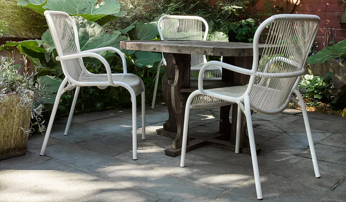 Vincent Sheppard Sitzmöbel in Weiß auf einer Terrasse mit Holztisch und Pflanzen im Hintergrund, stimmungsvolle Gartenatmosphäre.