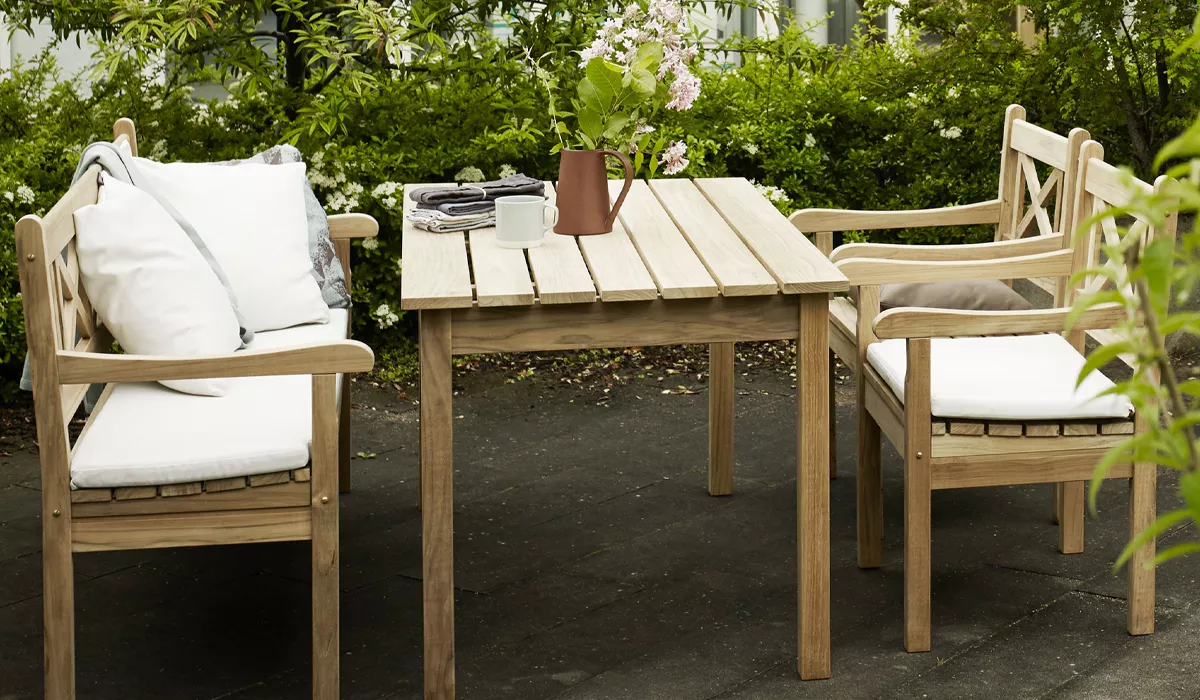Skagerak-Tisch und Stühle aus hellem Holz auf einer Terrasse mit grüner Hecke im Hintergrund, dekoriert mit Kissen und Blumen.