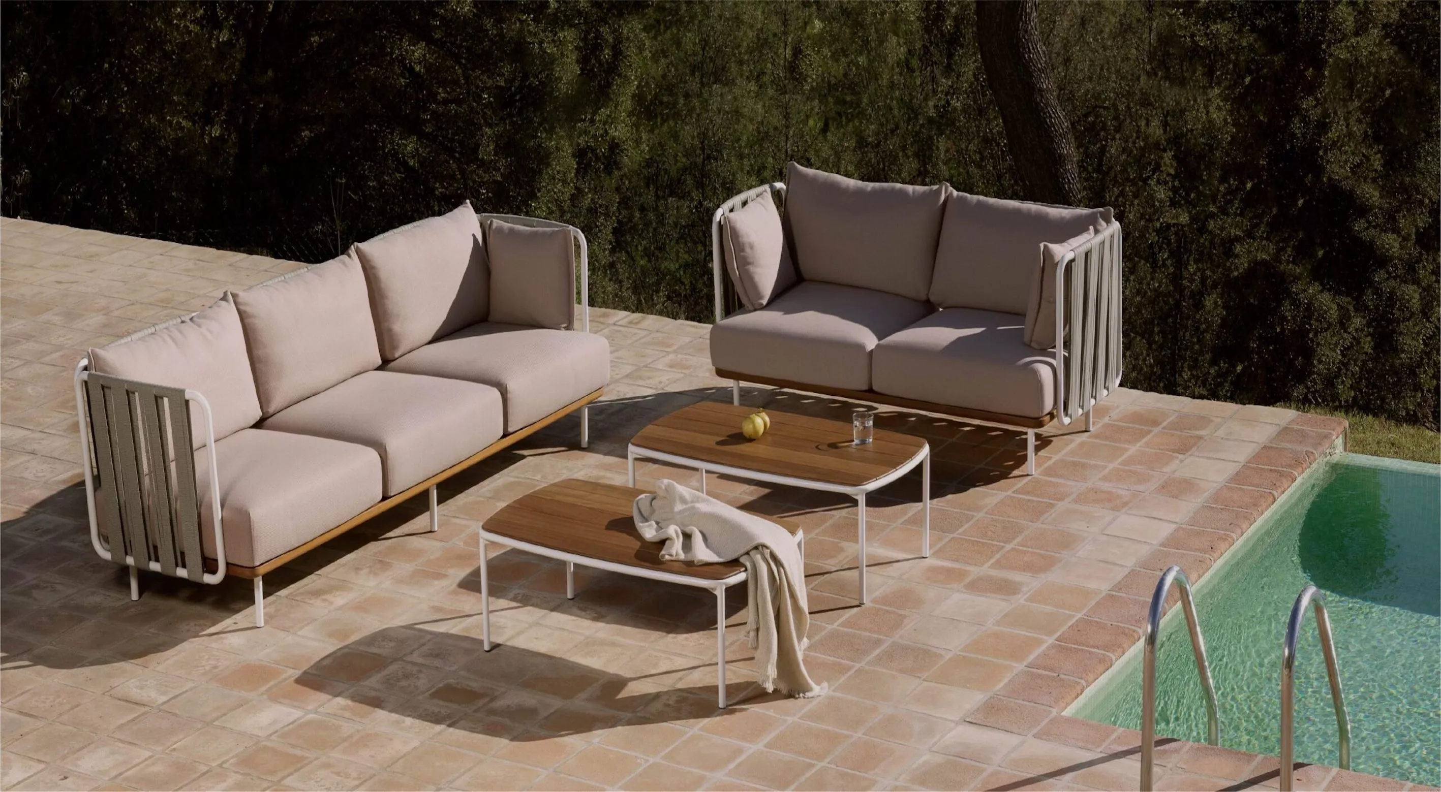 BIVAQ Teja Outdoor-Sofas in Beige auf einer Terrasse neben einem Pool, umgeben von Bäumen, mit zwei passenden Holztischen.