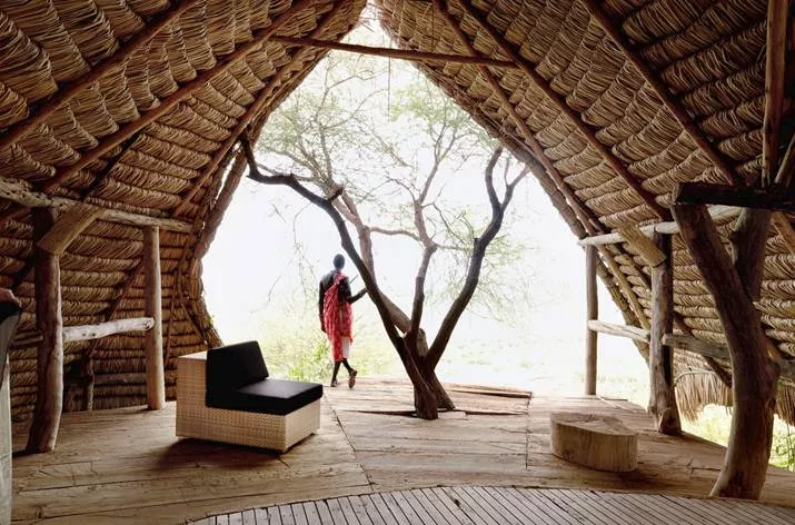 Eine Dedon Lounge Gartenmöbel steht in einer rustikalen Hütte mit Strohdach im Vordergrund während eine Person in traditioneller Kleidung nach draußen schaut