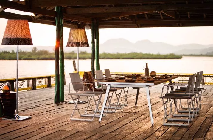 Dedon Seax Kollektion Seax Armlehnstuhl klappbar auf einer Terrasse mit Blick auf einen See bei Sonnenuntergang mit gedecktem Tisch und Lampen