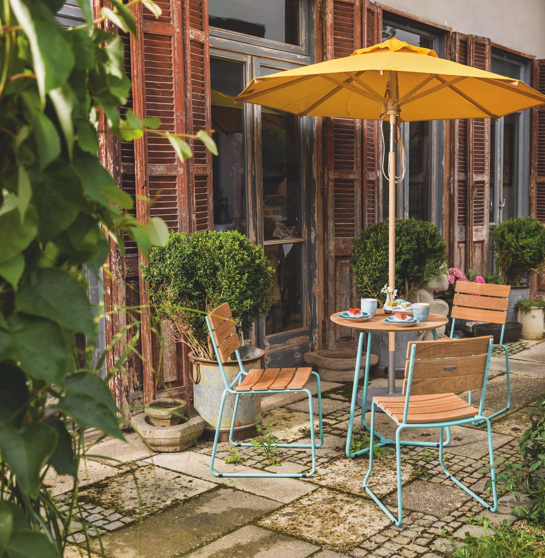 Weishäupl Balkon-Bistrotisch in Holzoptik mit gelbem Sonnenschirm, umgeben von Pflanzen auf einer rustikalen Terrasse.
