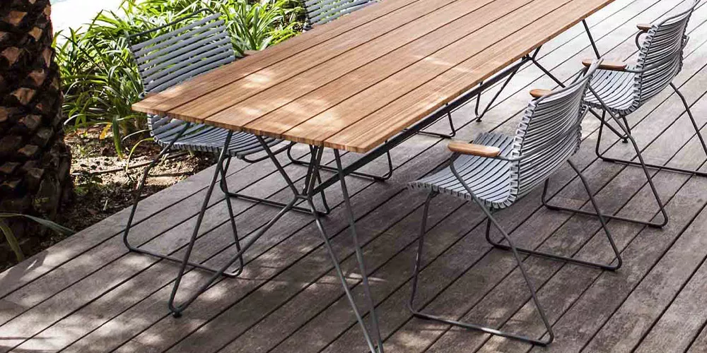 Houe Kollektion Sketch, Holztisch mit Metallgestell, umgeben von grauen Stühlen, auf einer Terrasse mit Pflanzen im Hintergrund.