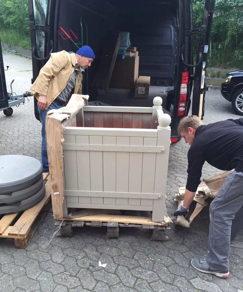 Zwei Männer entladen einen lackierten Orangerie Pflanzkübel aus Akazienholz aus einem Lieferwagen und entfernen die Verpackung.