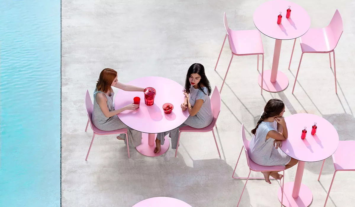 Drei Frauen sitzen an rosa Diabla Essplatzmöbeln auf einer Terrasse neben einem Pool, mit roten Gläsern und Flaschen auf den Tischen.