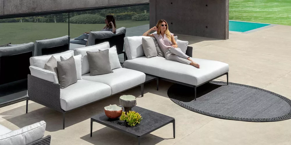 Frau entspannt auf einem modernen, weißen Tealenti Leaf Outdoor-Sofa mit grauen Kissen auf einer Terrasse neben einem Pool.