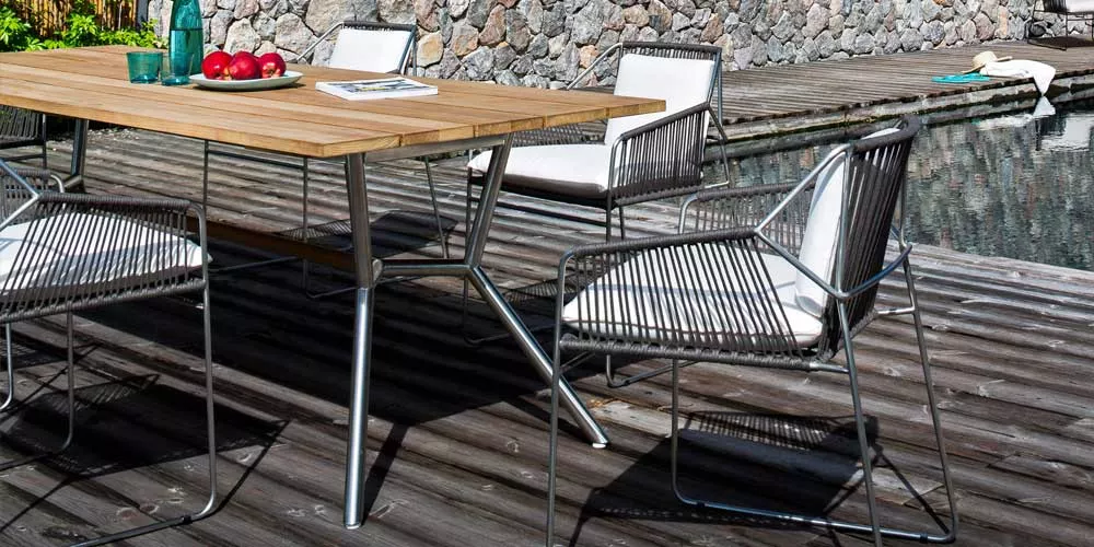 Oasiq Reef Essgruppe in Grau mit Holztisch und Stühlen auf Holzterrasse vor Steinmauer und Pool, sommerliche Gartenatmosphäre.