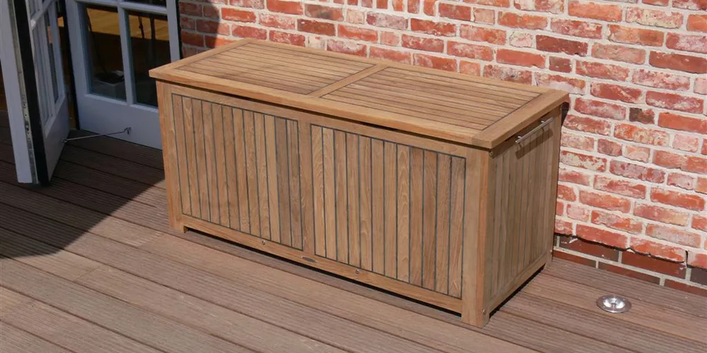 Royal Mirage Kissentruhe aus Holz in Naturfarbe auf einer Terrasse vor einer Ziegelwand, mit geschlossenem Deckel, stimmungsvolles Bild.