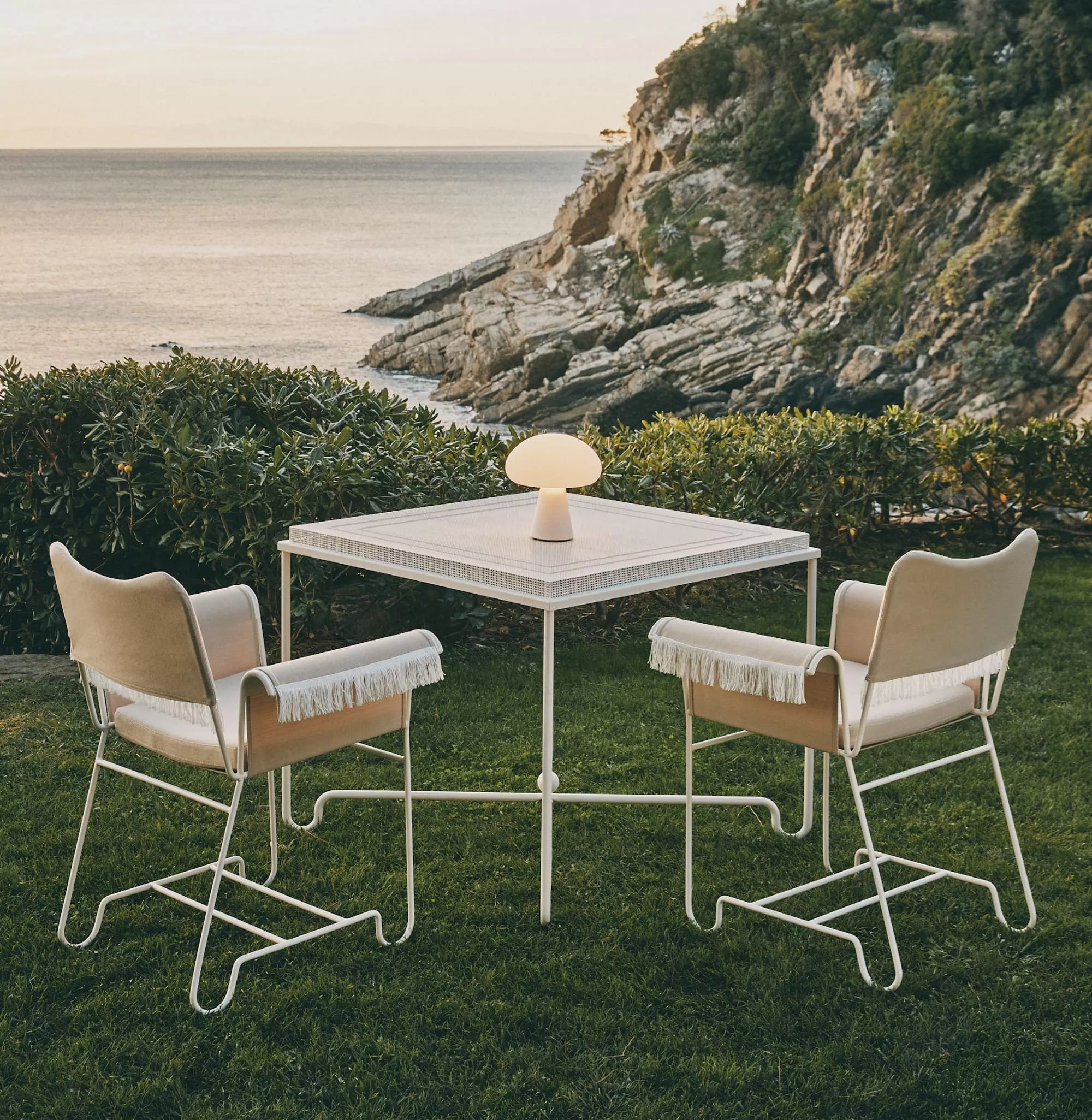 Zwei GUBI Tropique Stühle und ein Tisch mit Lampe stehen auf einer Wiese mit Blick auf das Meer und eine felsige Küste im Hintergrund