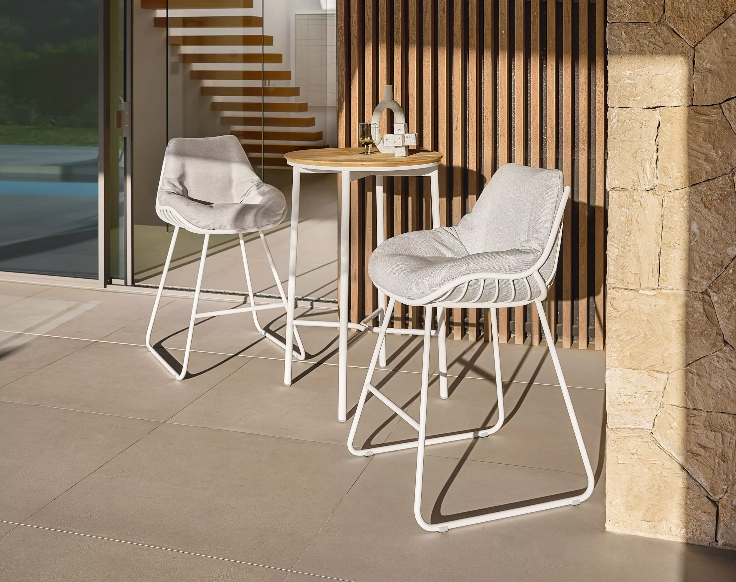 Zwei Solpuri Chill-A Barhocker mit weißen Gestellen und grauen Kissen stehen an einem runden Holztisch auf einer Terrasse bei Tageslicht.