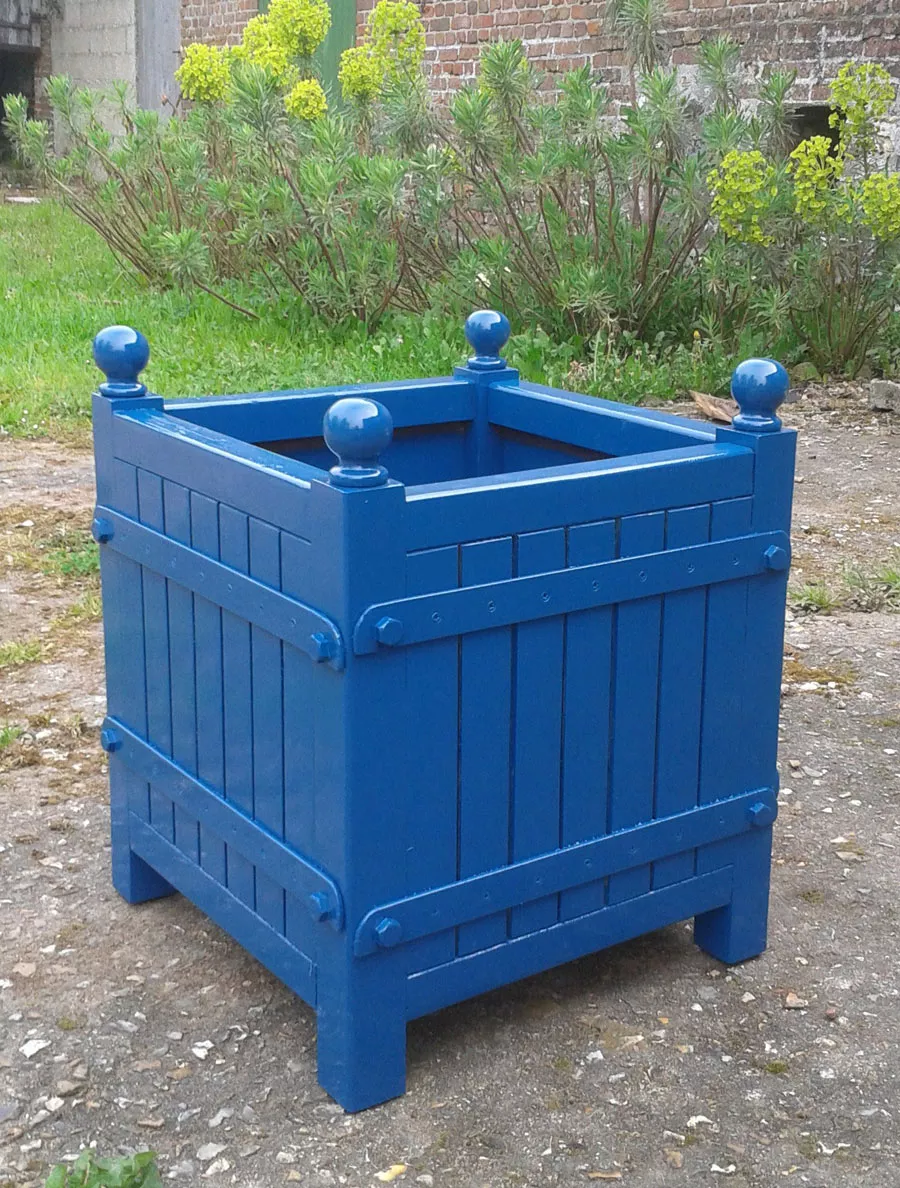 Blauer lackierter Orangerie Pflanzkübel aus Akazienholz steht auf einem gepflasterten Weg im Garten mit grünen Pflanzen im Hintergrund