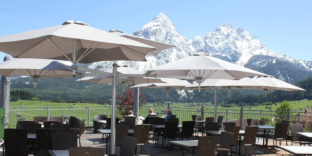 Paraflex Sonnenschirme in Beige auf einer Terrasse mit Bergblick, umgeben von Tischen und Stühlen, bei sonnigem Wetter.