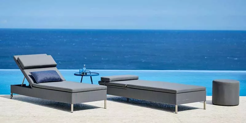 Zwei graue Cane-line Rest Sonnenliegen mit blauen Kissen am Pool vor dem Meer, begleitet von einem runden Beistelltisch und Hocker.