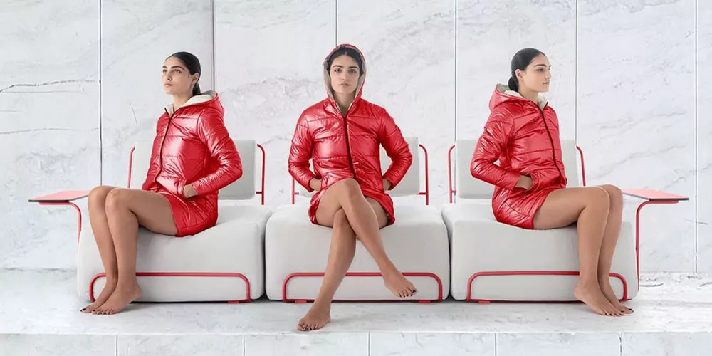 Drei Frauen in roten Diabla Lilly Daunenjacken sitzen auf weißen, modernen Bänken vor einer Marmorwand.