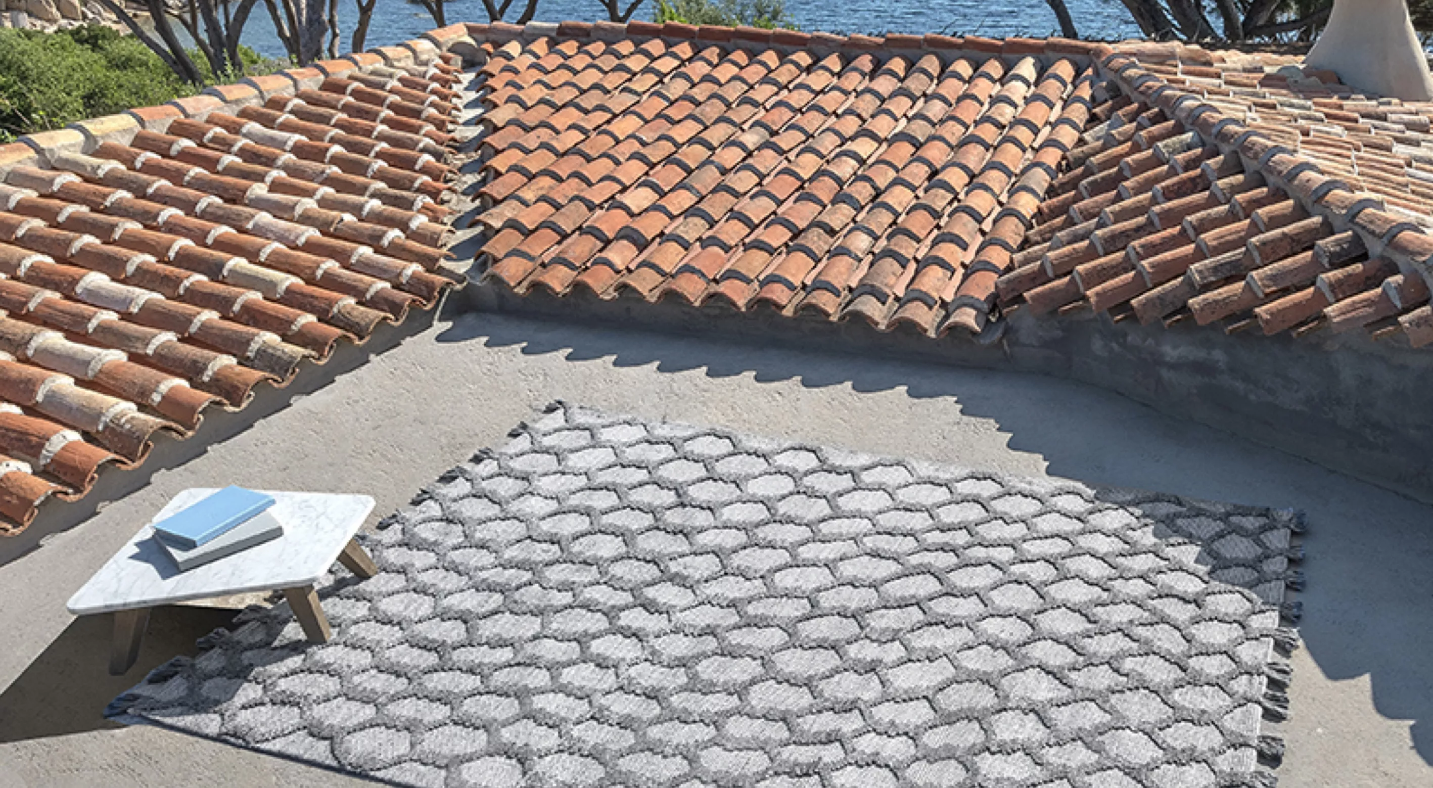 Grauer Outdoor-Teppich von Villa Schmidt auf einer Dachterrasse mit Meerblick, neben einem kleinen Tisch mit Büchern, in sonniger Atmosphäre.
