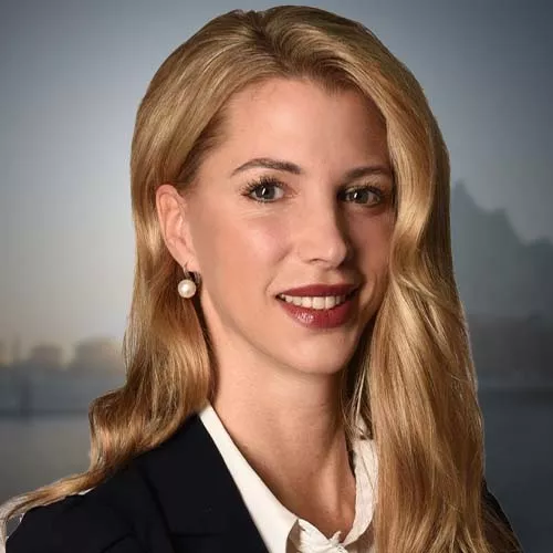 Eine lächelnde Frau mit blonden Haaren in einem schwarzen Blazer vor einem unscharfen Hintergrund, der eine Stadtlandschaft zeigt.