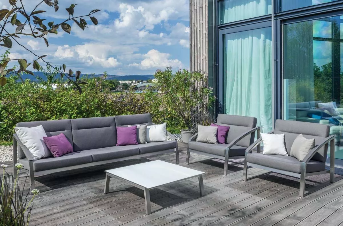 Grau-weiße Todus Alcedo Loungegruppe auf Holzterrasse vor modernem Gebäude mit Glasfront und grüner Gartenkulisse.