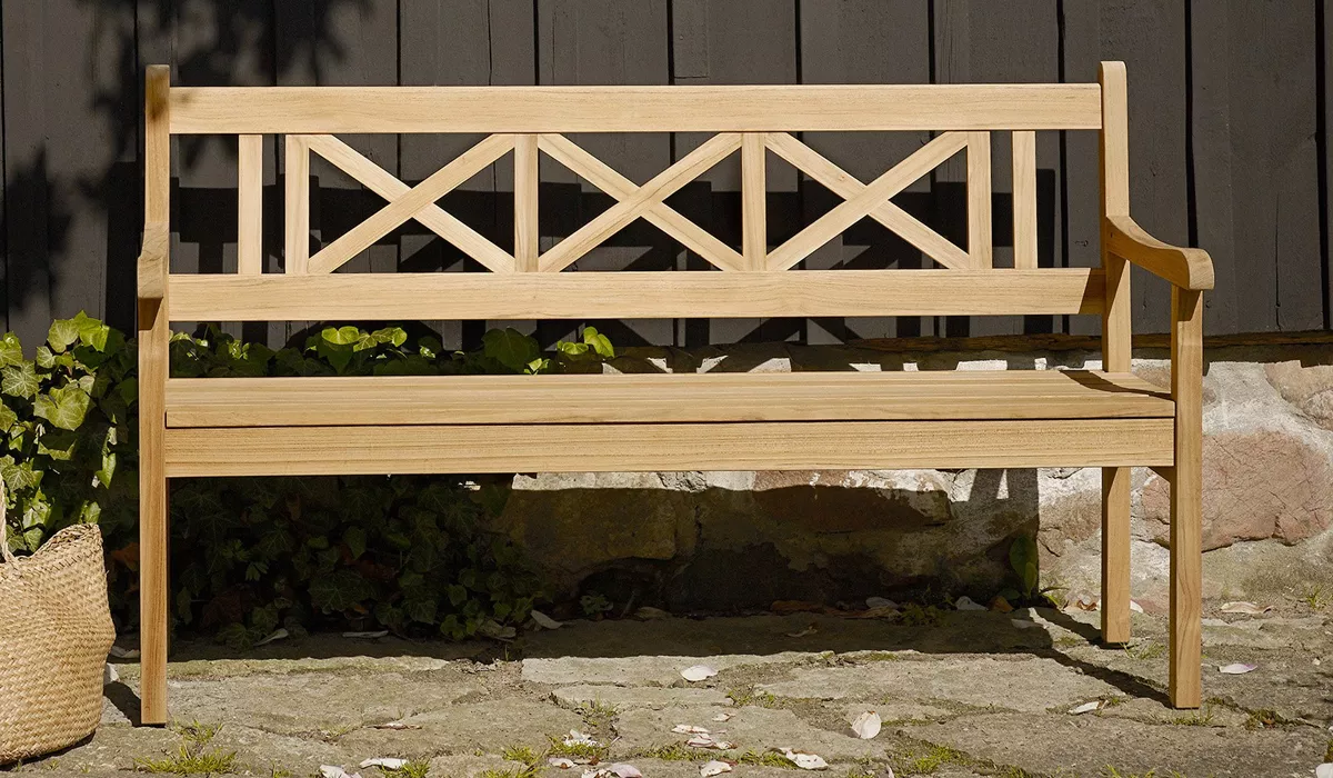 Skagerak Bank aus hellem Holz vor einer dunklen Holzwand, umgeben von Pflanzen, in einem sonnigen Garten.