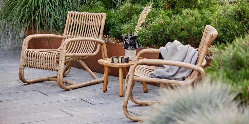 Cane-line Curve Loungegruppe in Naturfarbe auf einer Terrasse mit Pflanzen im Hintergrund, bestehend aus zwei Sesseln und einem kleinen Tisch.