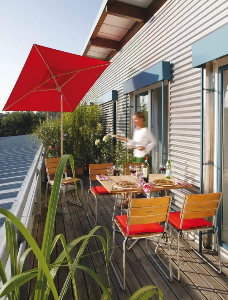 Klapptisch Balcony von Weish in Rot auf einer Terrasse mit Holzstühlen und rotem Sonnenschirm, stimmungsvolles Outdoor-Setting.