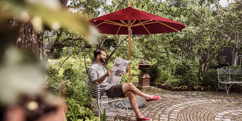 Ein Mann sitzt unter einem roten Glatz Piazzino Sonnenschirm im Garten und liest eine Zeitung.