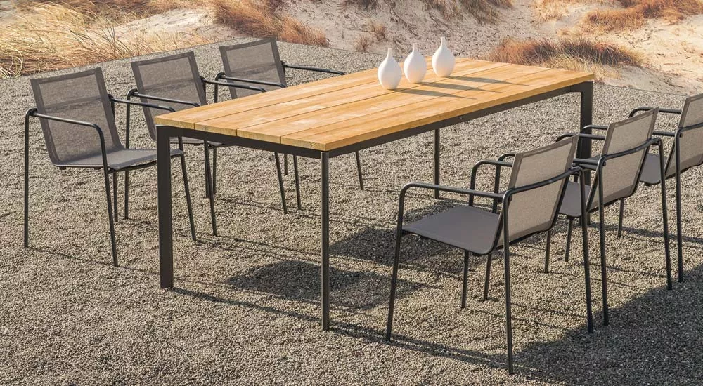 Fischer Möbel Taku Essgruppe in Schwarz mit Holztisch und acht Stühlen auf Kiesboden vor einer Dünenlandschaft.