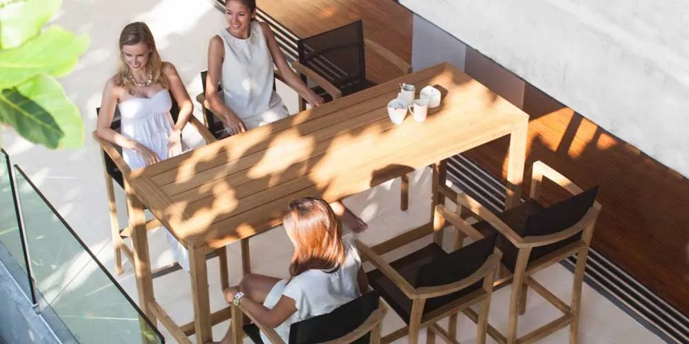 Drei Frauen sitzen an einem Royal Botania Red Label XQI Essgruppe Gartentisch aus Holz, umgeben von natürlichem Licht auf einer Terrasse.