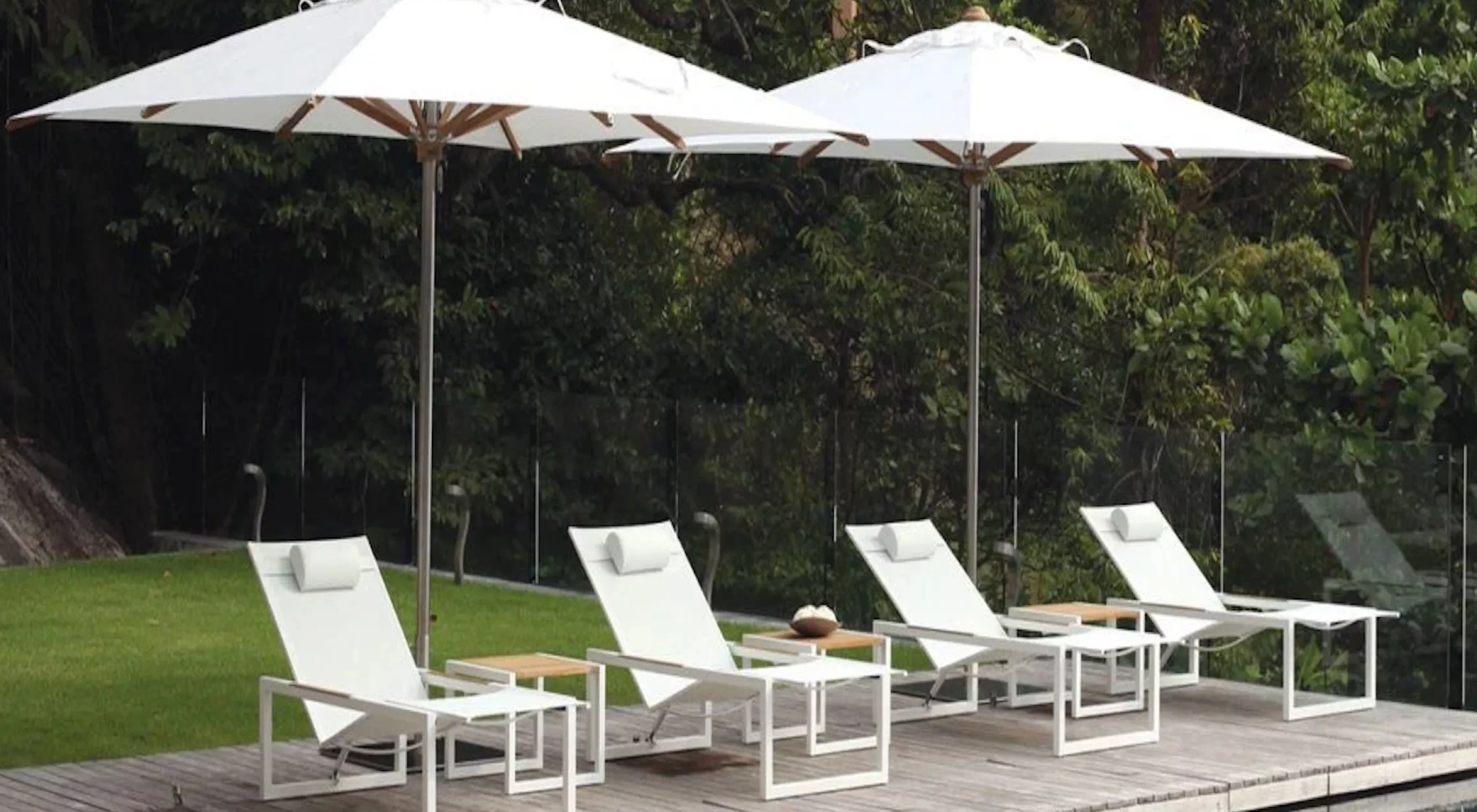 Vier weiße Sonnenliegen und Sonnenschirme der Marke Villa Schmidt auf einer Holzterrasse vor einem grünen Garten.