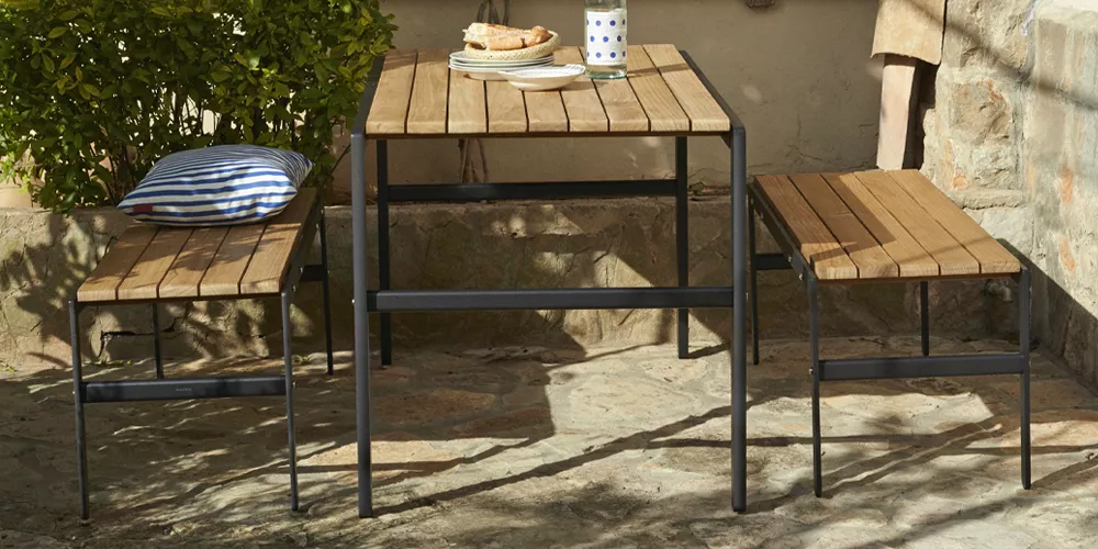 Skagerak Reform Set aus hellem Holz und schwarzem Metall, bestehend aus einem Tisch und zwei Bänken, auf einer sonnigen Terrasse.