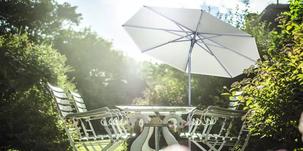 Glatz Alu-Smart Sonnenschirm in Weiß, aufgestellt in einem sonnigen Garten mit grüner Hecke und Gartenmöbeln im Hintergrund.