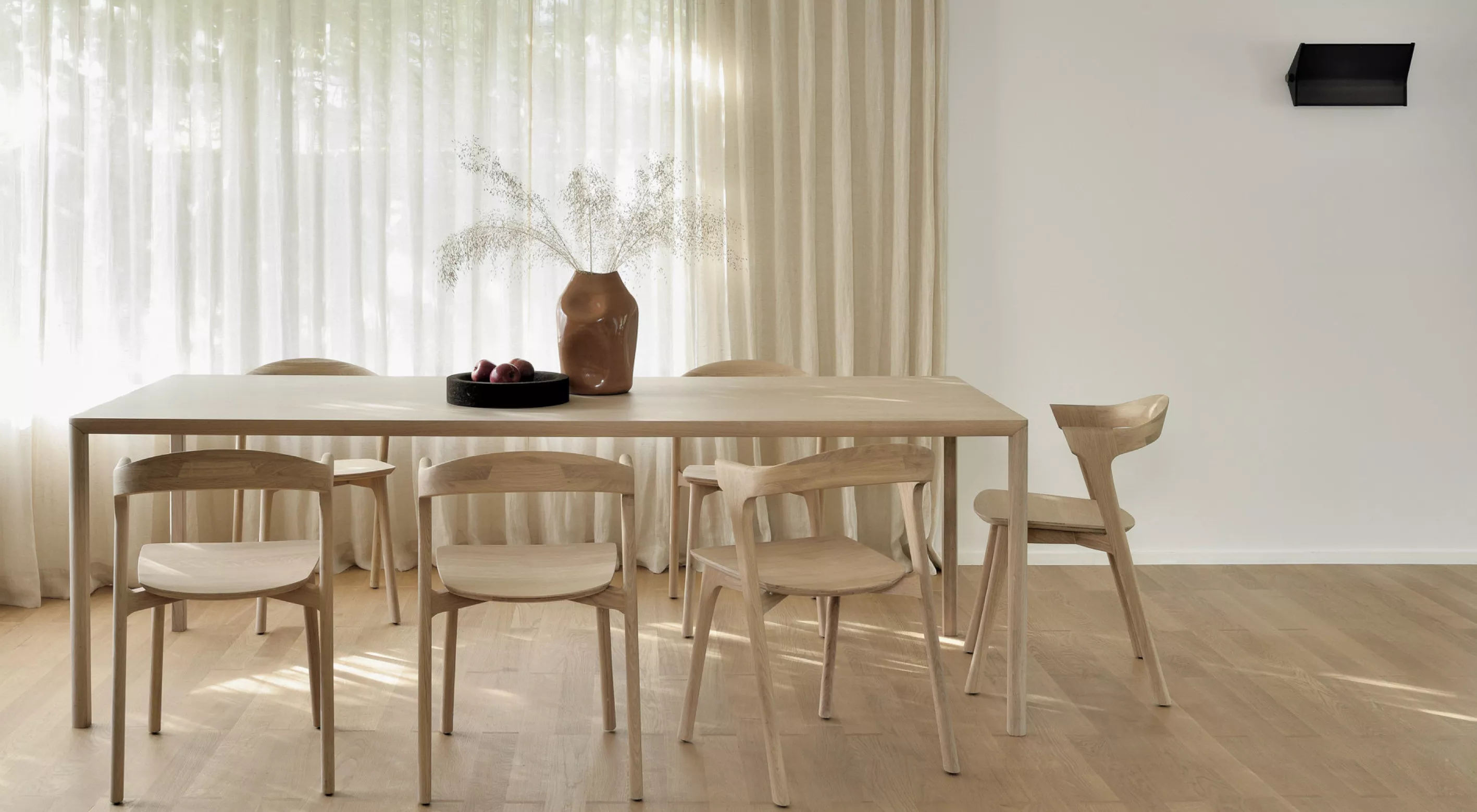 Oak Air Esstisch und Oak Bok Stühle in hellem Holz vor einem Fenster mit weißen Vorhängen, dekoriert mit einer Vase und Obstschale.
