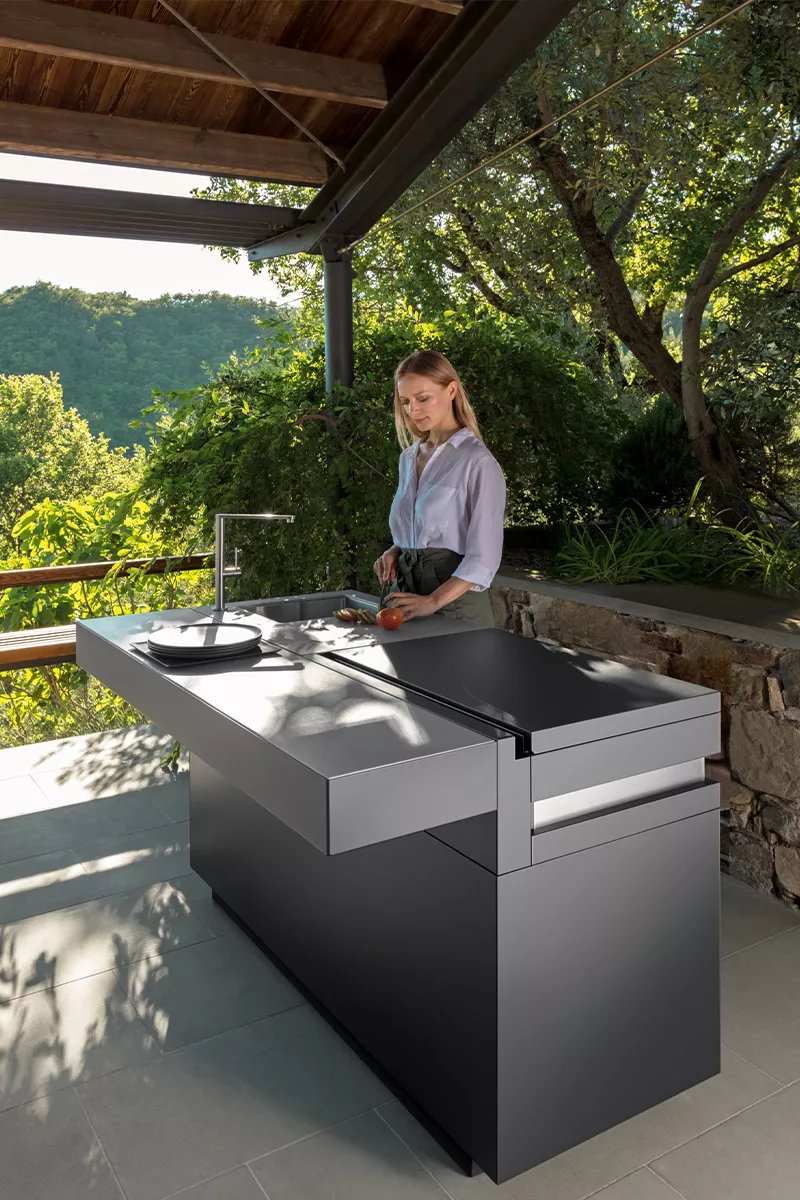 Frau bereitet Essen an der grauen Tikal Talenti Outdoor-Küche auf einer Terrasse mit Blick auf grüne Landschaft zu.