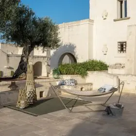Talenti Panama Hängematte mit Gestell in Beige auf einer sonnigen Terrasse vor einer historischen Steinmauer.