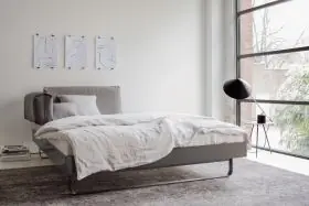 Graues NOVA Bett von Villa Schmidt in modernem Schlafzimmer mit großen Fenstern und minimalistischer Dekoration, seitliche Perspektive.