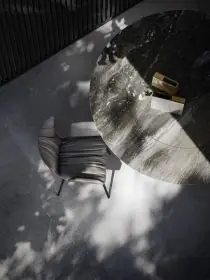 Ein Tadao III Stuhl von Draenert steht neben einem runden Marmortisch auf einem grauen Boden in einem stimmungsvollen Bild mit Licht und Schatten.