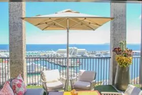 Cape Umbrellas PREMIUM St. James Sonnenschirm in Beige auf einer Terrasse mit Meerblick, umgeben von gemütlichen Sitzmöbeln und Blumen.