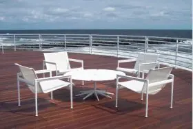 Vier weiße Knoll Studio 1966 Collection Lounge Sessel und ein weißer Tisch stehen auf einer Holzterrasse mit Blick auf das Meer bei bewölktem Himmel.