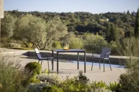 Todus Alca Esstisch in Schwarz mit HPL-Tischplatte, umgeben von zwei Stühlen, auf einer Terrasse mit Blick auf einen Garten.