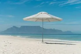 CapeUmbrellas ELITE Constantia Sonnenschirm in Weiß auf einem sandigen Strand mit Blick auf das Meer und einen Berg im Hintergrund.