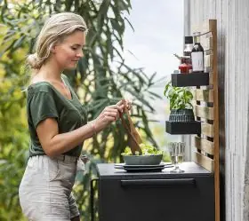 Frau kocht an der Cane-line Drop Outdoor-Küche in Schwarz mit 3 Regalen, im Garten vor einer Betonwand.
