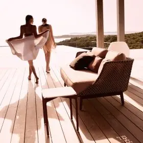 Zwei Frauen spazieren auf einer Terrasse am Meer neben einem Dedon Mbarq 3er-Sofa mit Kissen und einem Beistelltisch in einer stimmungsvollen Szene.