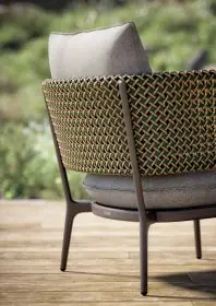 DEDON BELLMONDE Sessel mit geflochtenem Design und grauem Kissen steht auf einer Holzterrasse im Freien umgeben von grüner Natur