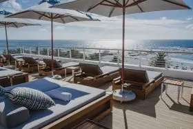 Cape Umbrellas PREMIUM Montague in Braun auf einer sonnigen Terrasse mit Meerblick und Liegestühlen, stimmungsvolle Atmosphäre.