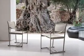 Zwei B&B Italia Mirto Outdoor Gartenstühle stehen auf einer Terrasse vor einem großen Baum und einer Steinmauer im Hintergrund.