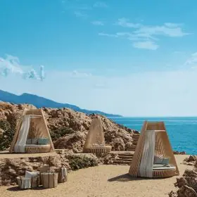 Ethimo Hut Loungebett in Naturtönen auf sandigem Strand mit Meerblick und Felsen im Hintergrund, stimmungsvolle Outdoor-Szene.