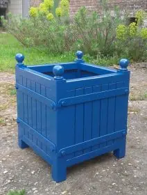 Blauer lackierter Orangerie Pflanzkübel aus Akazienholz steht auf einem gepflasterten Weg im Garten mit grünen Pflanzen im Hintergrund